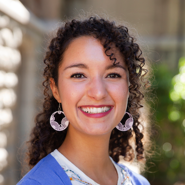 A woman smiling with long curly brown hair, wearing a purple cardigan and large lavender earrings.