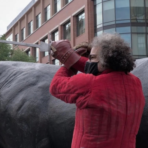 Torso shot of a curly-haired, male-presenting person, in profile, wearing a red  jacket, thick gloves, glasses, and black facemask, and wielding a longsword in a high guard. In the background is a statue of a bull and, further back, skyscrapers.