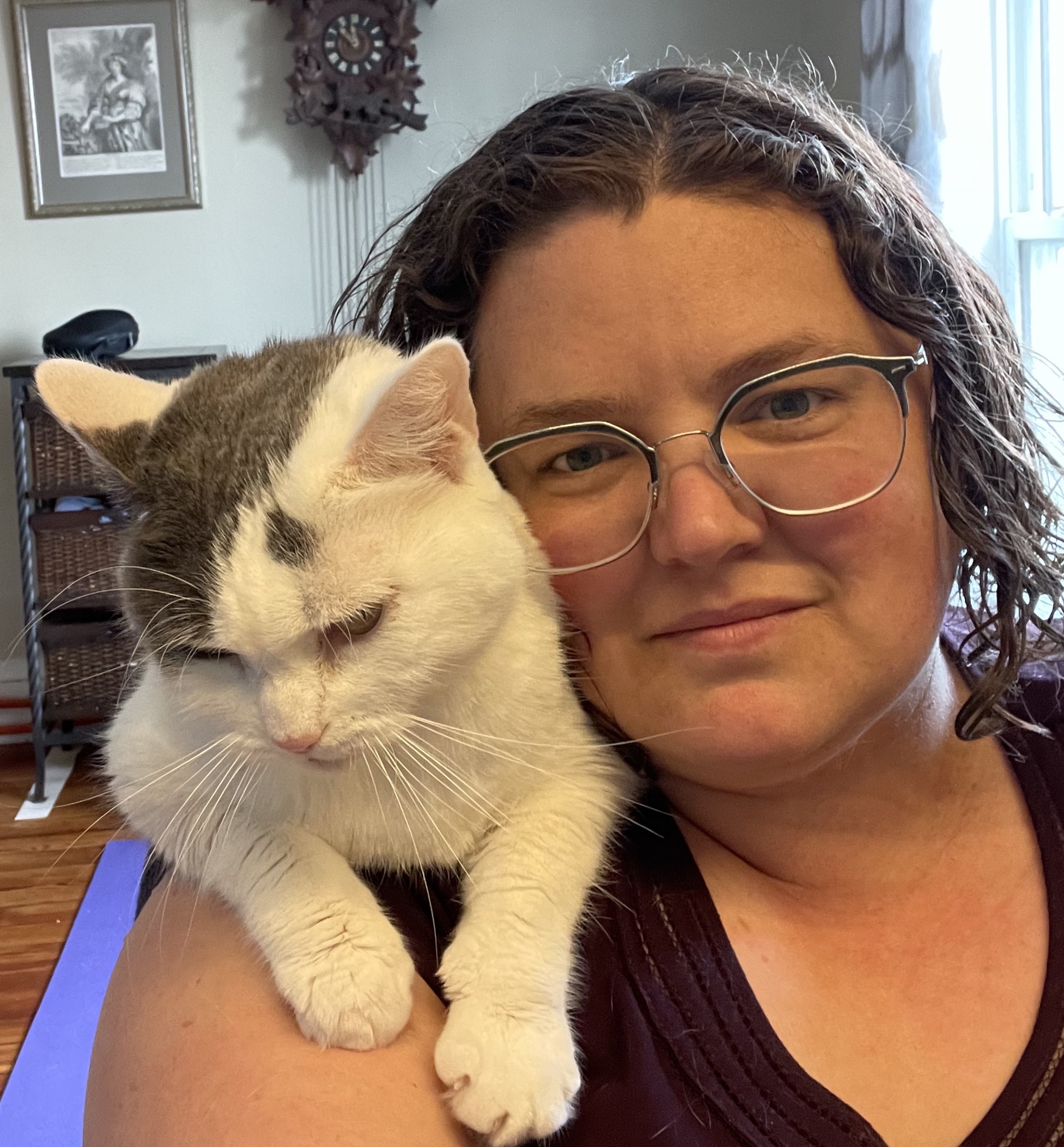 Andromeda looks straight at the camera. She's a white woman with glasses, wavy brown hair, and a half-smile. A white-and-grey cat perches on her shoulder, unimpressed.