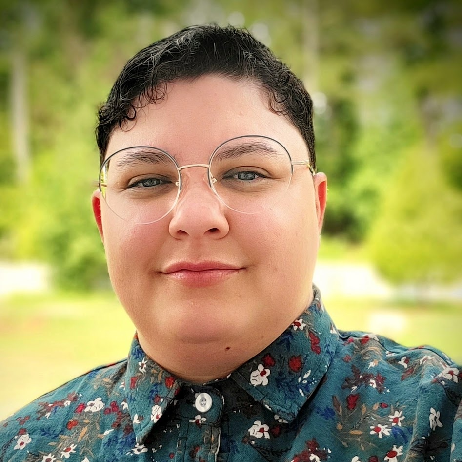 A light-complexion masculine-presenting individual with a short, curly haircut wearing round glasses and blue-green floral print buttoned down shirt.