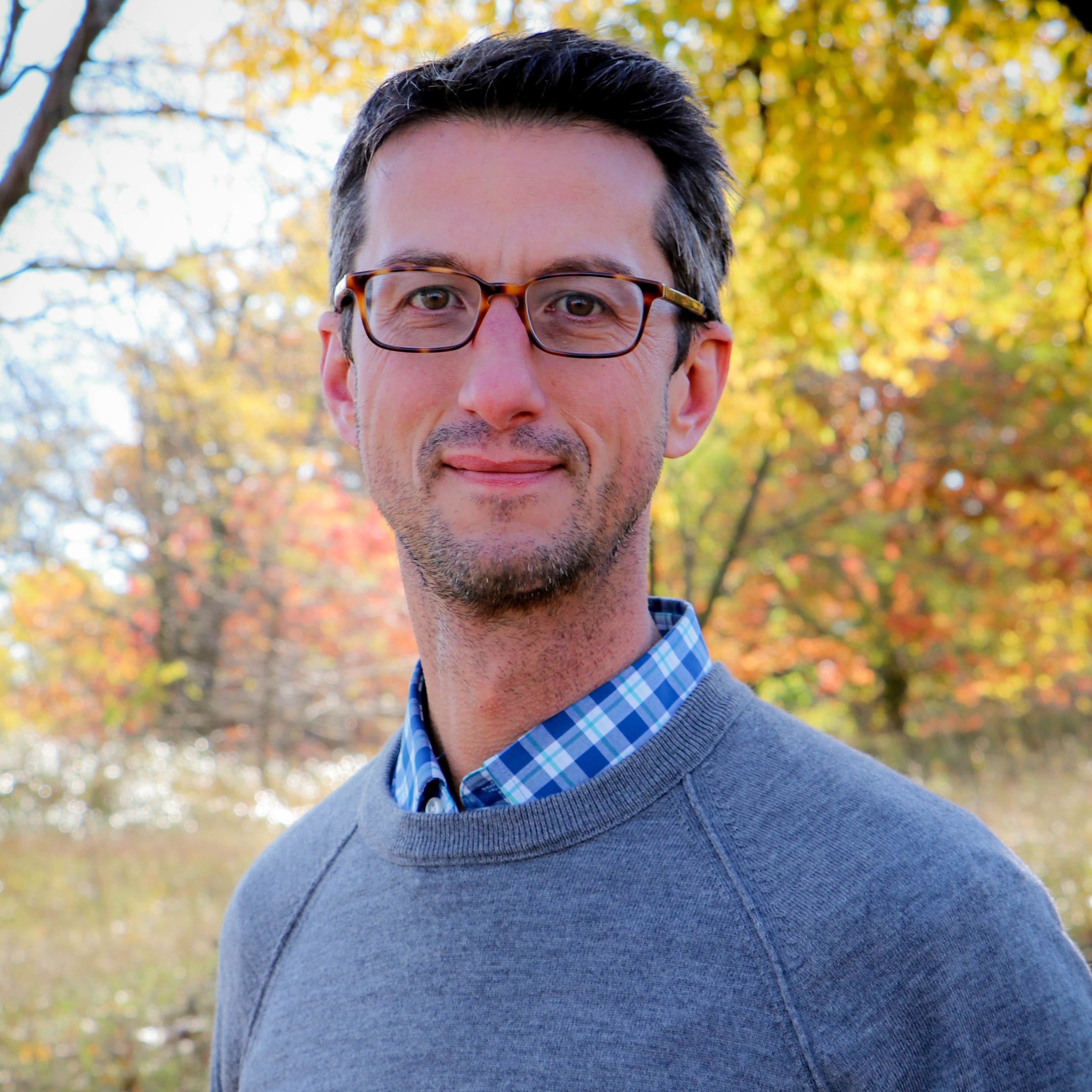 David in glasses and smiling stands against fall foliage in a grey sweater.