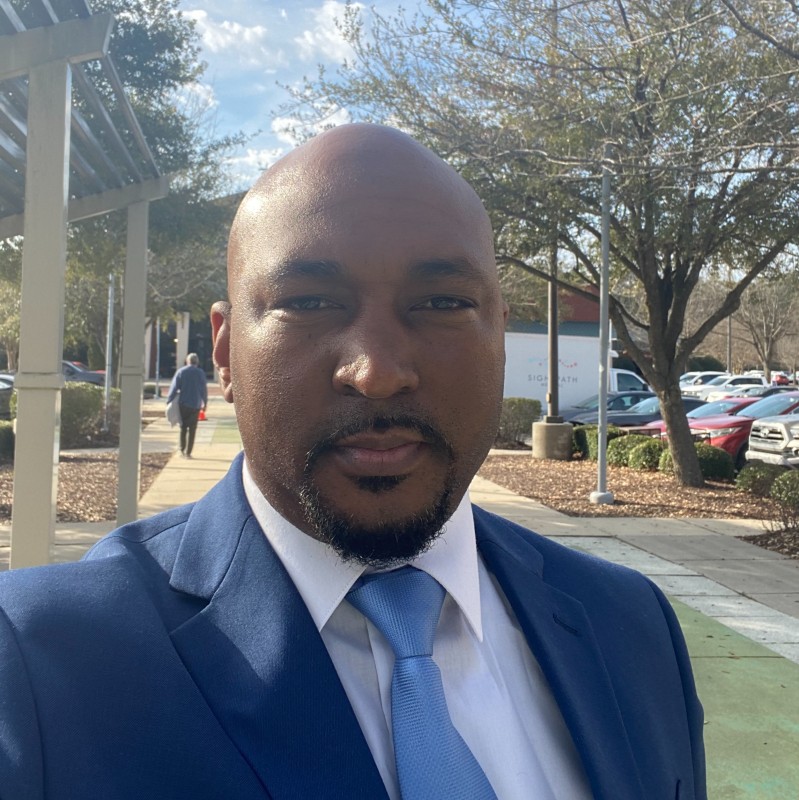 A bald headed black male with thinly trimmed mustache with hints of black and grey hair. Wearing a blue suit, white shirt and a light blue tie looking dapper yet professional with the car park in the background