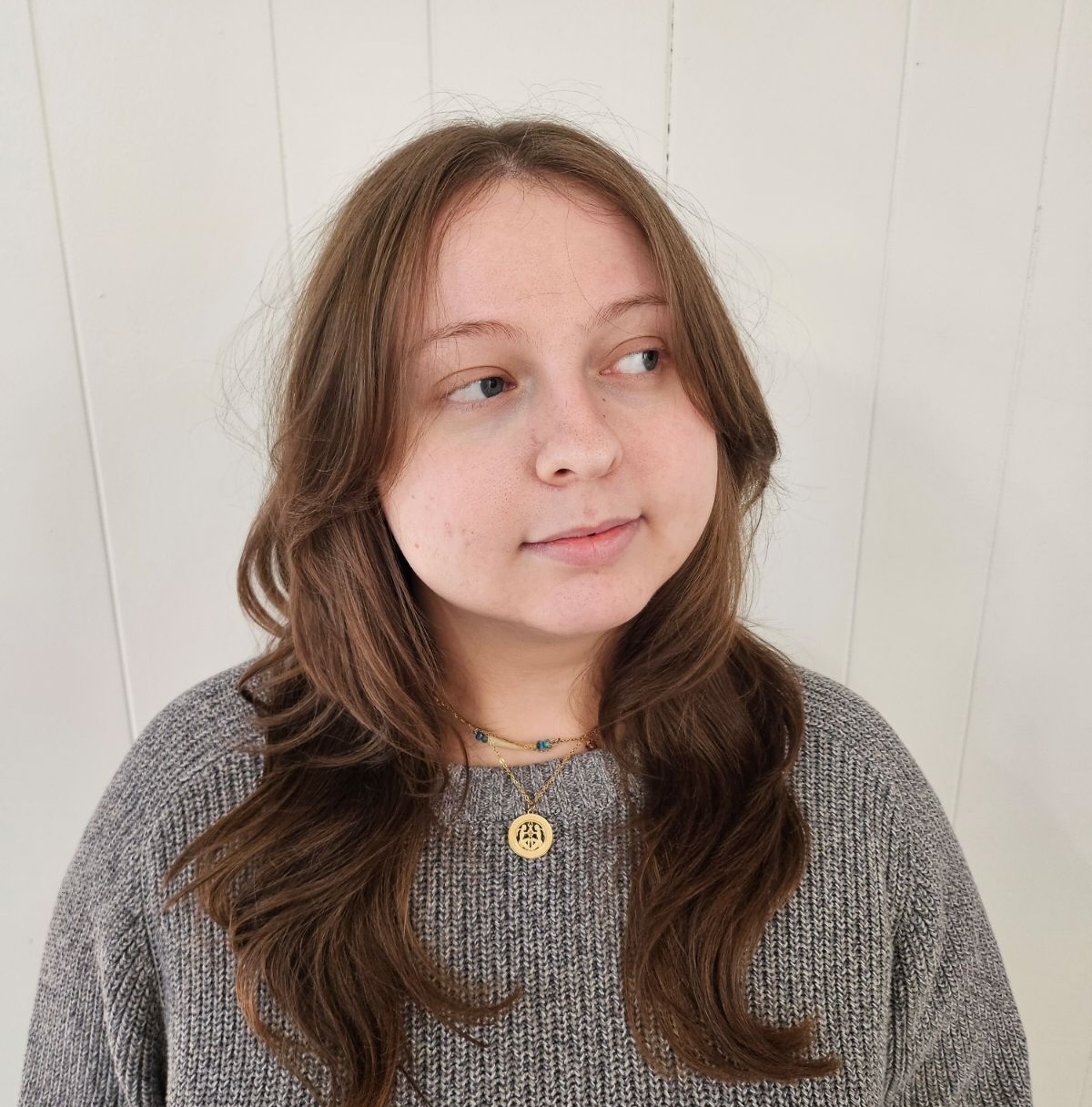 A color photograph of a non-binary individual from the waist up. The person is wearing a thick sweater, two necklaces made of dentalium shell, gold metal, and beads, and has their long hair down. The figure is looking off to the right side, smiling mildly. It is equal measures polite interest and disintrest.