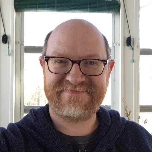 A bald, middle-aged white man with a close-trimmed red beard which is graying at the chin, wearing tortoiseshell glasses and a dark blue hoodie.