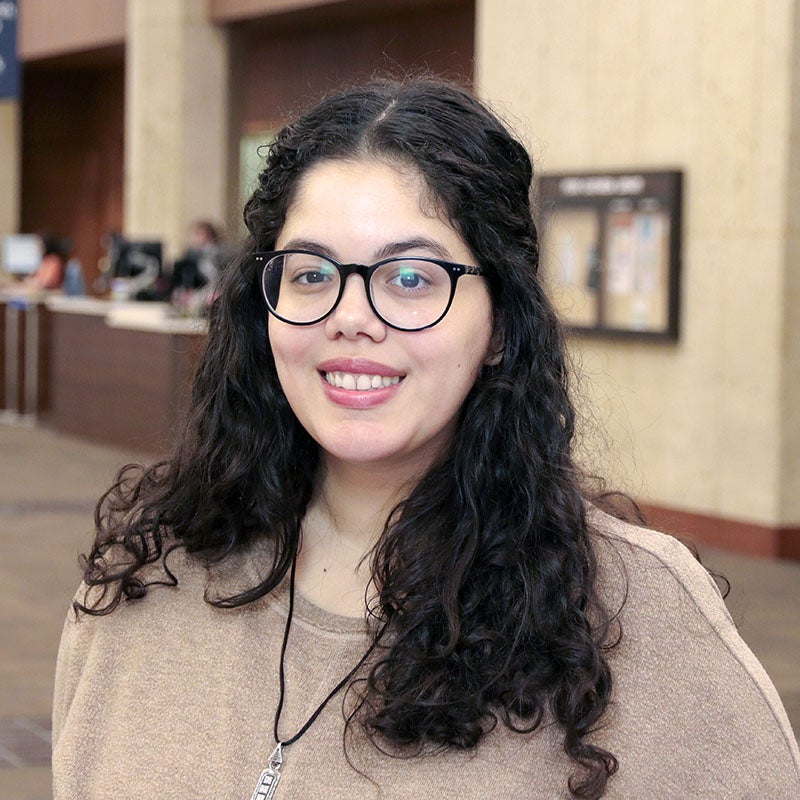 A young white latina woman with dark curly hair wearing dark-framed glasses is smiling at the camera. A library help desk is behind her.