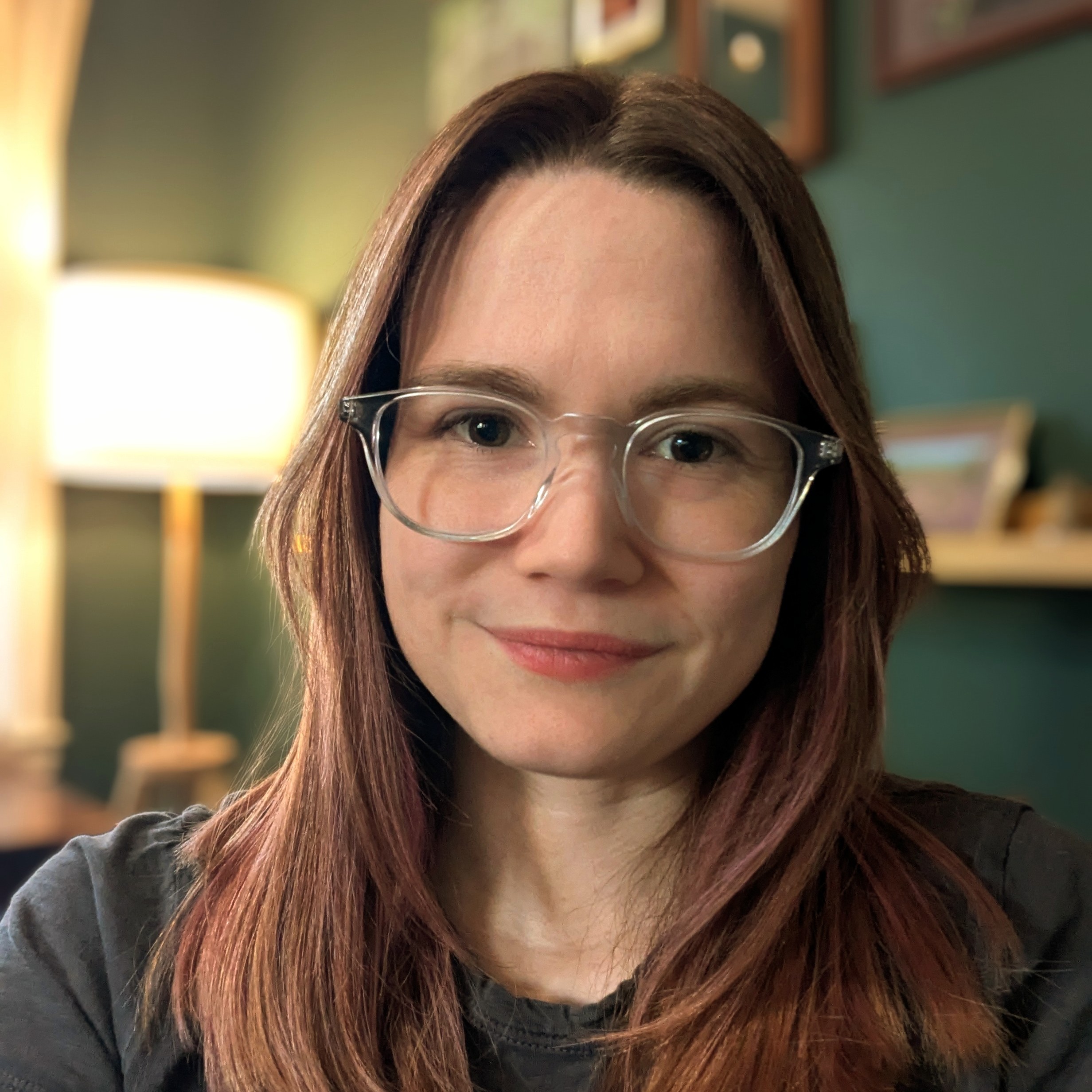 A white woman in her thirties with blond pink hair and glasses is smiling at the camera. A dark green wall with photos and a lamp is behind her.