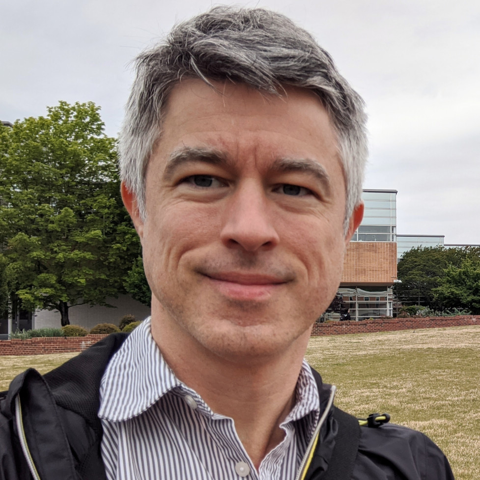 Selfie photograph of a middle-aged white man with graying hair, wearing a casual, striped shirt and a light nylon windbreaker. He is standing outside, in a field, with red brick university buildings behind him.