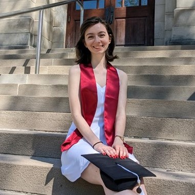 A young woman grins at the camera with her graduation cap and stole. She is posed on a staircase outside a university building.