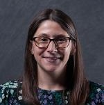 A slightly freckled white woman with brown hair wearing rounded brown eyeglasses, looking happy and professional, and wearing a black top with purple, green, and white flowers. There is a dark gray backdrop behind her.