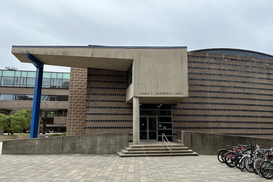 An exterior view of McDonnell Hall, a red and brown striped brick building with an angular overhang. Steps lead to the main entrance, which is flanked by an access ramp, bicycles, and stairs to the lower level.